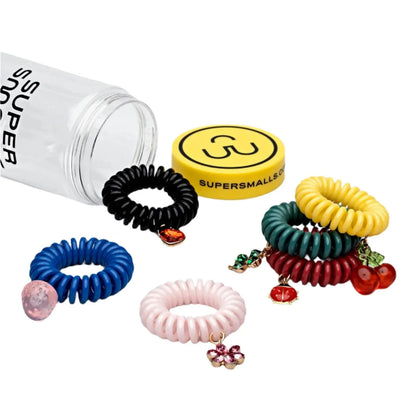 A tipped clear container showcasing an array of Super Smalls Charmed Life Hair Ties, each featuring colorful coiled designs adorned with jeweled charms in fruit and floral motifs. The container lid is yellow and branded with "Super Smalls," making it the ideal summer accessory.