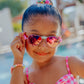 A young girl stands by the pool, smiling and holding her Bling2O Girls’ Hampton Beach Sunglasses with UV-protected lenses. She wears a pink checkered swimsuit, along with a beaded bracelet and a colorful hair tie.