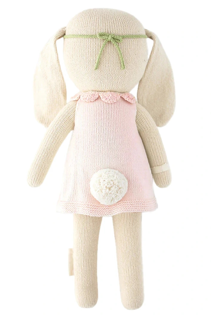 Hannah the Bunny - Little 13" by Cuddle + Kind is a knitted plush toy with long floppy ears, a pale pink dress, green headband, and white tail—photographed from the back. This adorable bunny makes an inspirational birthday gift.