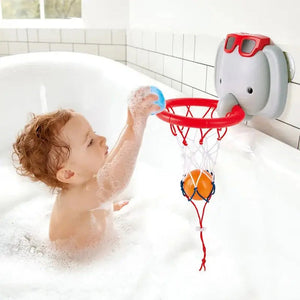 A young child enjoys bath time fun with the Hape Bath Basketball Elephant Pal, aiming a blue ball at the red hoop attached to the tub wall. An orange ball already swishes in the net while the cheerful elephant pal bath toy floats nearby, making playtime even more delightful.