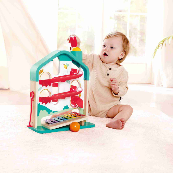 A toddler sits on a light carpet in a bright room, playing with the Hape Melody Mansion Ball Run—a colorful, house-shaped wooden toy featuring ball ramps and a built-in xylophone board.