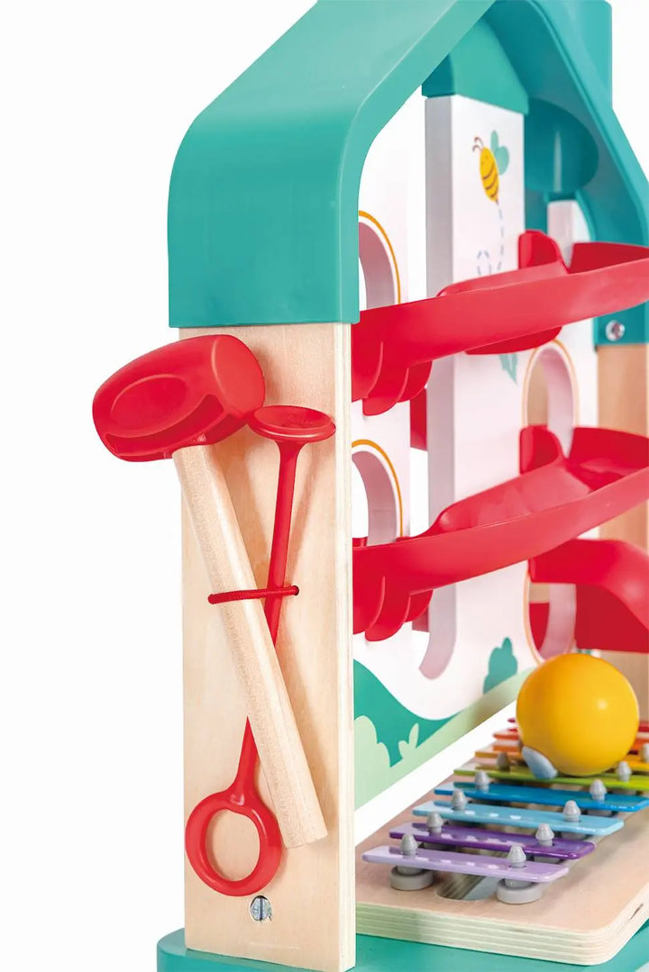 Close-up of the Hape Melody Mansion Ball Run, a house-shaped toy featuring a xylophone board, colorful ramps, a yellow ball, and two red mallets attached to the side with a red band.