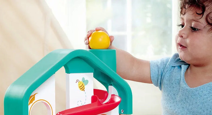 A young child places a yellow ball at the top of the colorful Hape Melody Mansion Ball Run, which features red and green components, indoors.