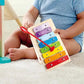 A baby sits on a carpeted floor holding the Hape Melody Mansion Ball Run, with wooden toys, a yellow ball, and a house-shaped toy nearby.