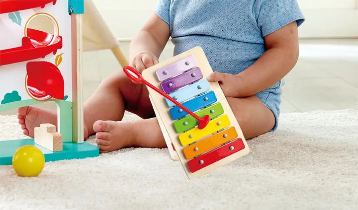 A baby sits on a carpeted floor holding the Hape Melody Mansion Ball Run, with wooden toys, a yellow ball, and a house-shaped toy nearby.