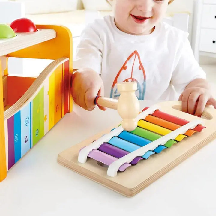 A child sits at a table indoors, wearing a white shirt with an orange design, and plays with the Hape Pound & Tap Musical Bench using a small mallet. This colorful musical toy encourages creativity and helps develop hand-eye coordination.