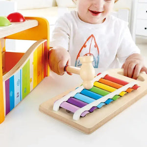 A child sits at a table indoors, wearing a white shirt with an orange design, and plays with the Hape Pound & Tap Musical Bench using a small mallet. This colorful musical toy encourages creativity and helps develop hand-eye coordination.