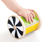 A child’s hand touches the Hape Spinning Drum w/ Mirror, a wooden developmental baby toy featuring a black-and-white spiral design on one end and a green and yellow body, placed on a white surface—perfect for sensory play.