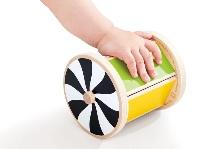 A child’s hand touches the Hape Spinning Drum w/ Mirror, a wooden developmental baby toy featuring a black-and-white spiral design on one end and a green and yellow body, placed on a white surface—perfect for sensory play.