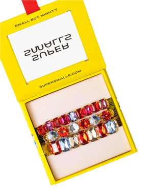 A yellow box labeled "Super Smalls" holds the Happy Hour Bracelet Set—three colorful, stretchy, stackable kids' bracelets with oversized gemstones side by side, perfect for playful jewelry lovers.