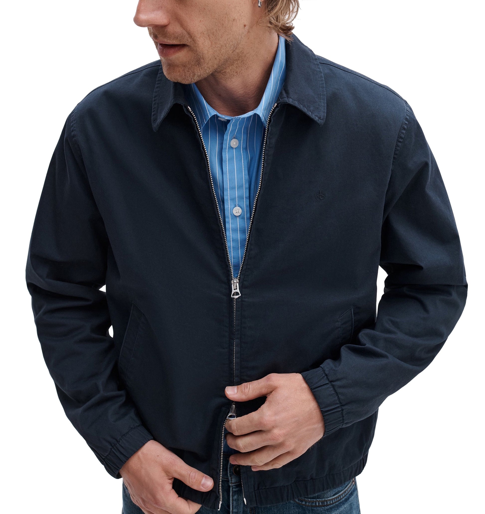 A man in a Rag & Bone Harrington Cotton Jacket over a blue striped button-up shirt stands against a white background, adjusting his jacket—a modern take on classic British sportswear.