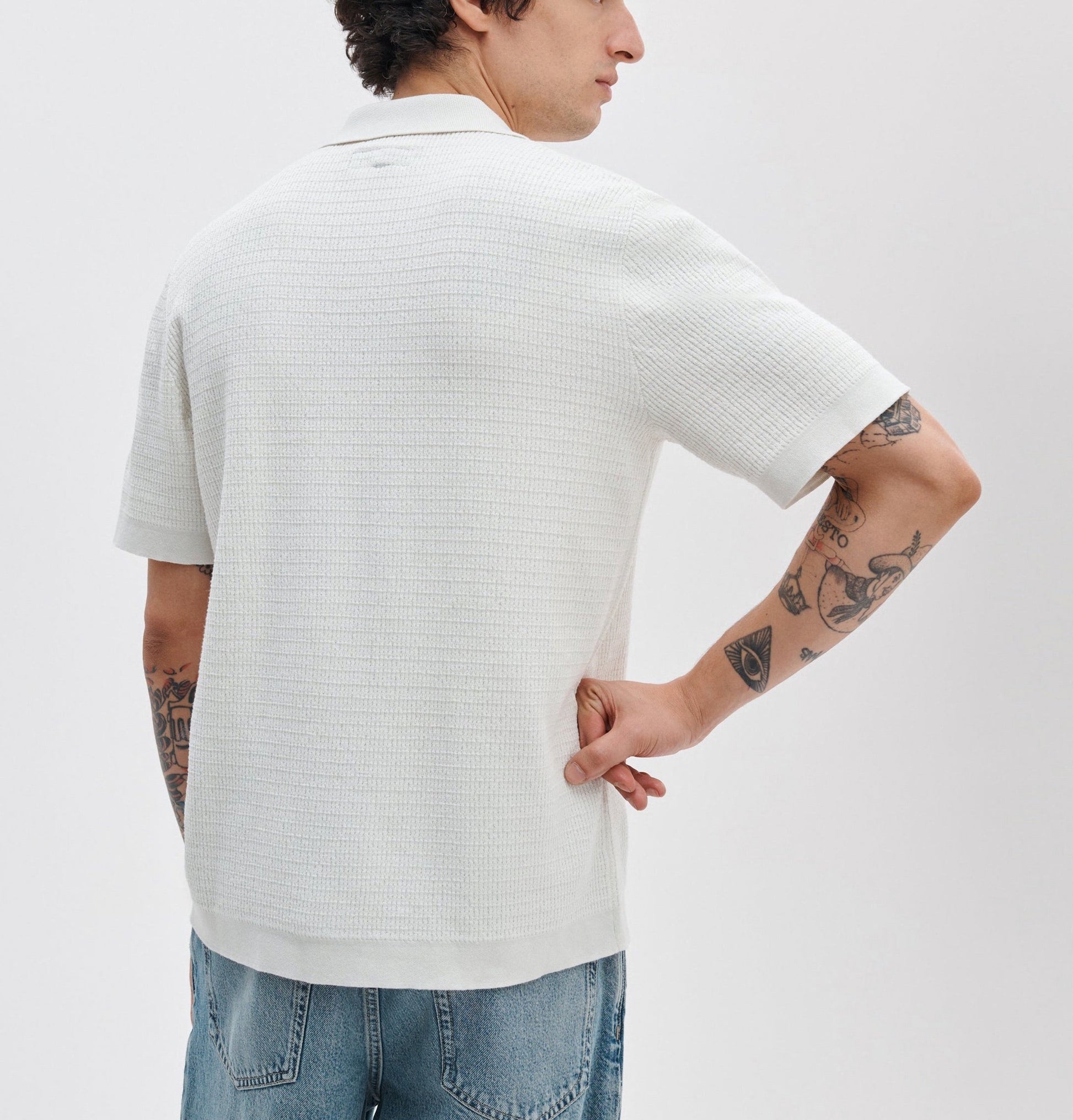 A person faces away from the camera with one hand behind their back, showing tattoos on both arms while wearing the Rag & Bone Harvey Camp Shirt, a retro-inspired white knit with subtle stripes, and light blue jeans.