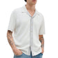 A person wears the Rag & Bone Harvey Camp Shirt, a retro-inspired white short-sleeve button-up with a subtle striped effect, styled over a gray top and blue jeans; hands in pockets, tattoos visible on both arms.
