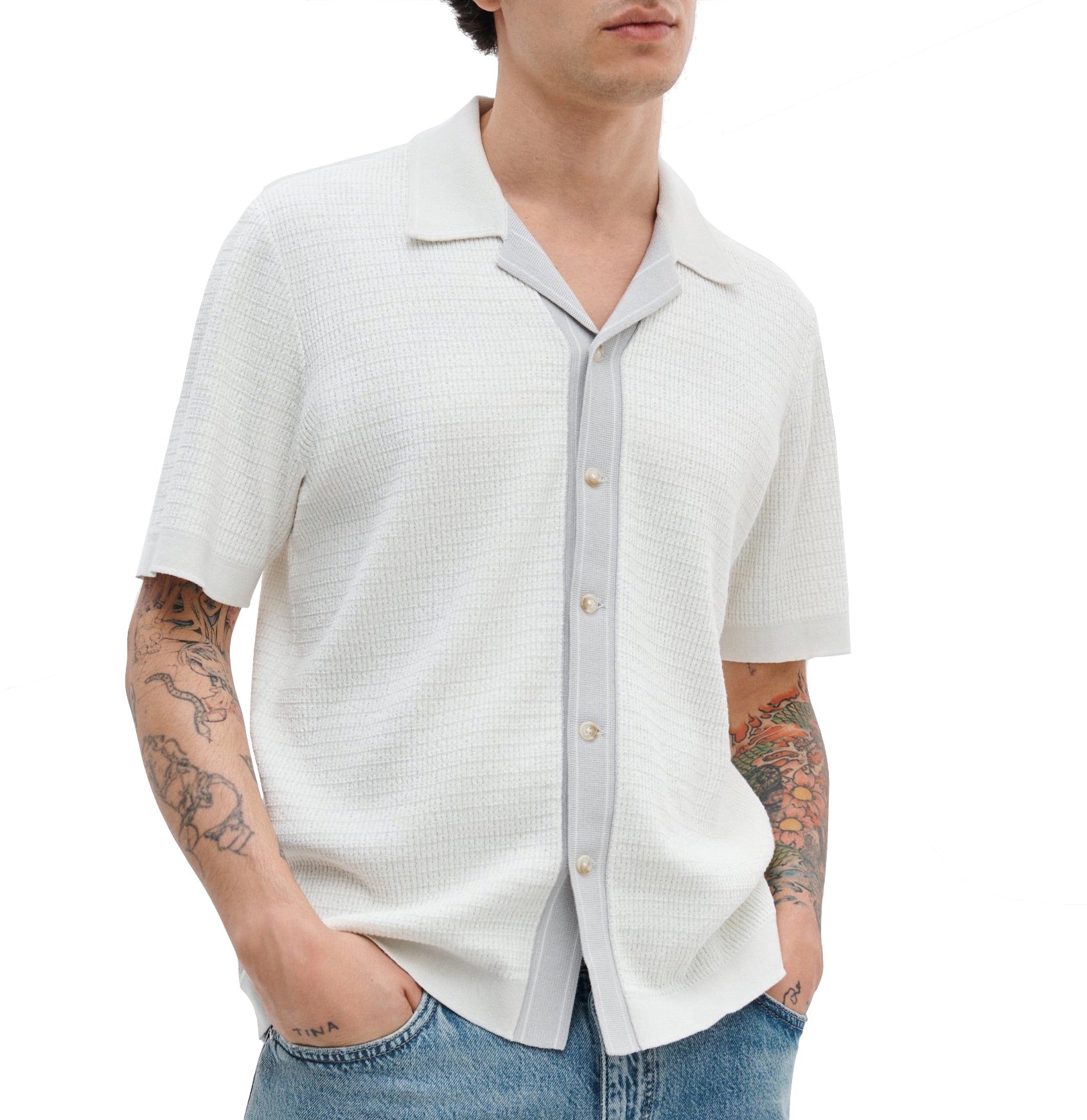 A person wears the Rag & Bone Harvey Camp Shirt, a retro-inspired white short-sleeve button-up with a subtle striped effect, styled over a gray top and blue jeans; hands in pockets, tattoos visible on both arms.