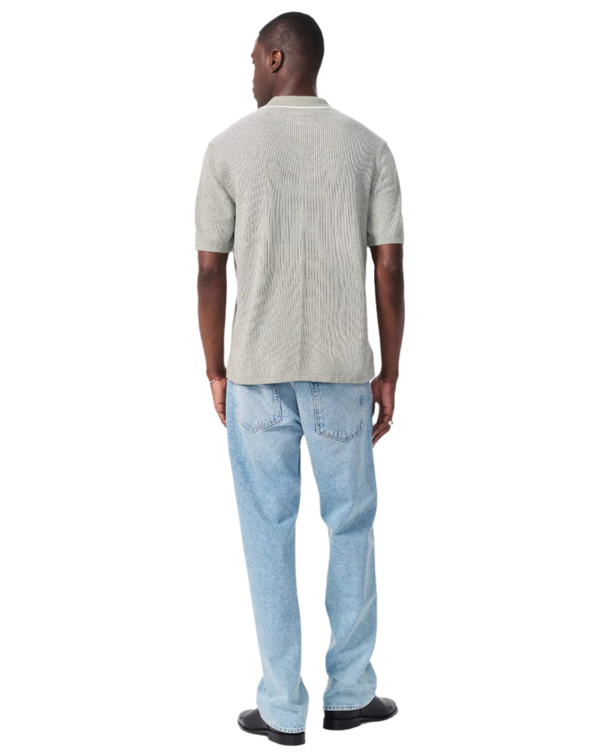 A man stands facing away, wearing the Rag & Bone Harvey Knit Polo Shirt in light gray, paired with light blue jeans and black shoes, against a white background.