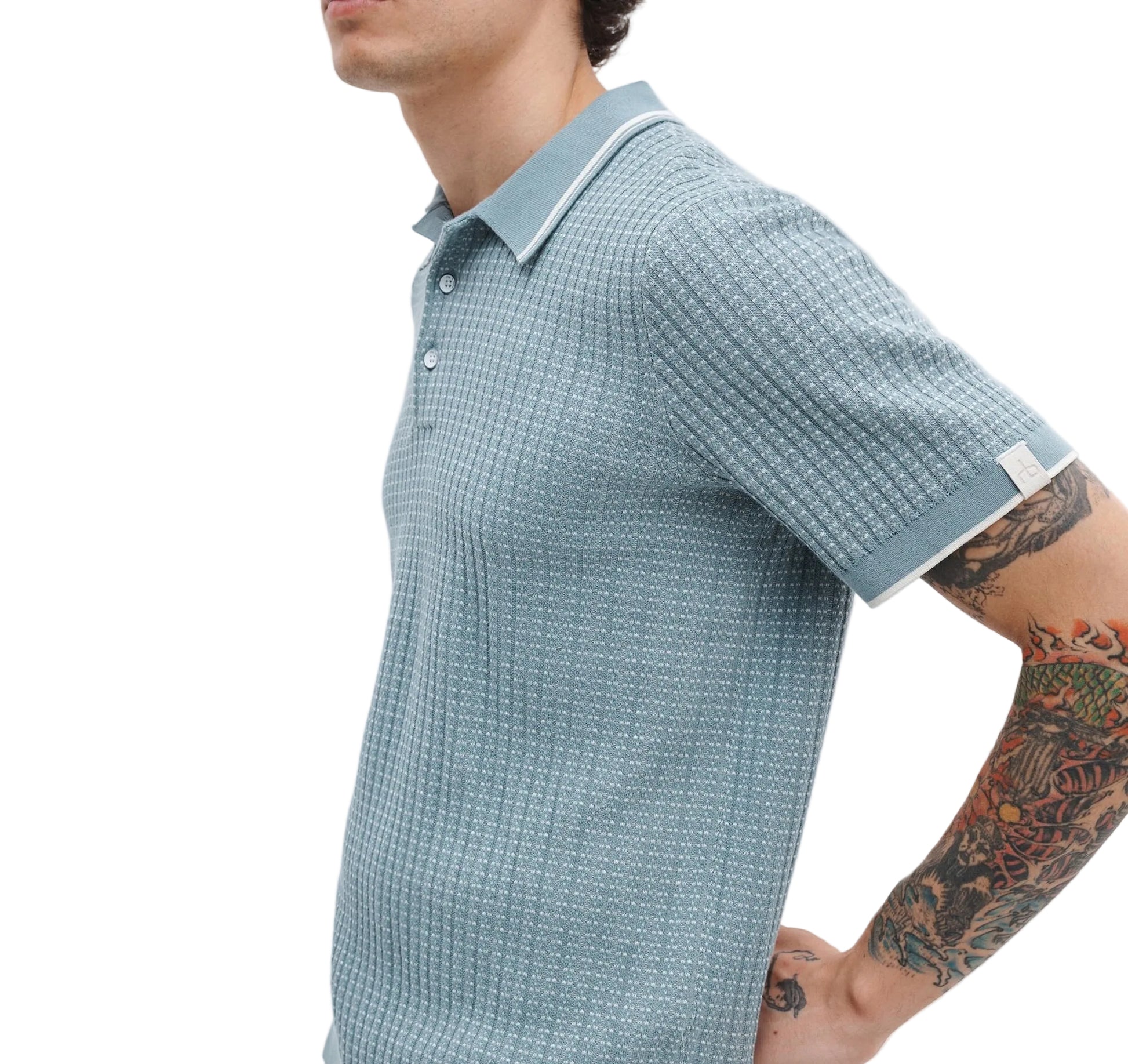 A man models the Rag & Bone Harvey Rib Polo, a light blue patterned cotton-blend short-sleeve shirt, revealing his tattooed right arm against a white backdrop for a subtle vintage feel.