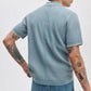 A person with tattooed arms is seen from the back against a plain background, wearing Rag & Bone’s Harvey Rib Polo in light blue and blue jeans.