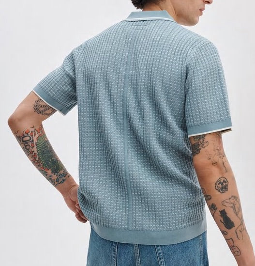 A person with tattooed arms is seen from the back against a plain background, wearing Rag & Bone’s Harvey Rib Polo in light blue and blue jeans.