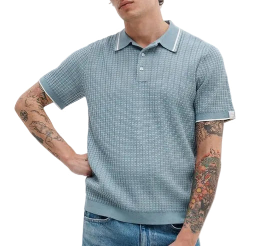 A person wearing the Rag & Bone Harvey Rib Polo—a retro-inspired light blue short-sleeve cotton-blend collared sweater—with jeans, showing tattooed arms.