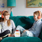 Two children sit on a teal couch wearing Yoto Headphones Orange with adjustable headbands. They are looking at each other while a small cube-like device with a pixelated screen sits between them. A lamp and artwork are in the background.