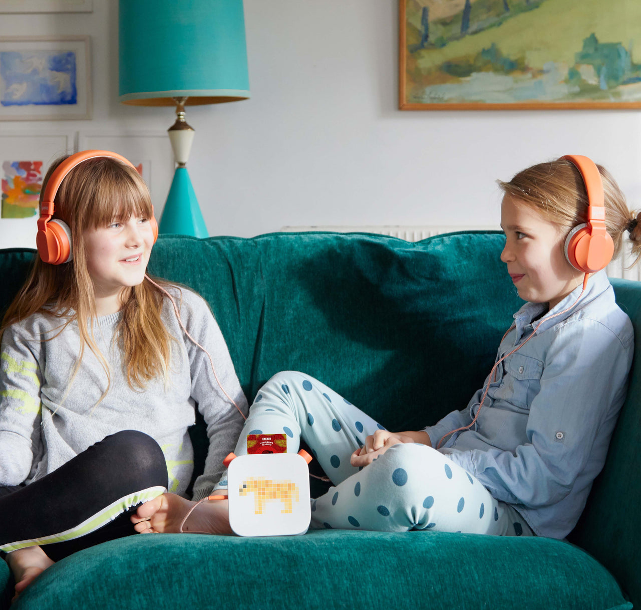Two children sit on a teal couch wearing Yoto Headphones Orange with adjustable headbands. They are looking at each other while a small cube-like device with a pixelated screen sits between them. A lamp and artwork are in the background.
