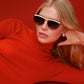 A woman with long blonde hair wears Krewe's Heidi Sunglasses, featuring an architectural metal brow bar, and an orange turtleneck, reclining against a solid red background.