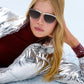 A woman with long blonde hair wears Krewe's Heidi Sunglasses—vintage-inspired with an architectural metal brow bar—along with a shiny silver jacket and red turtleneck, lying on snow and resting her head on one hand.