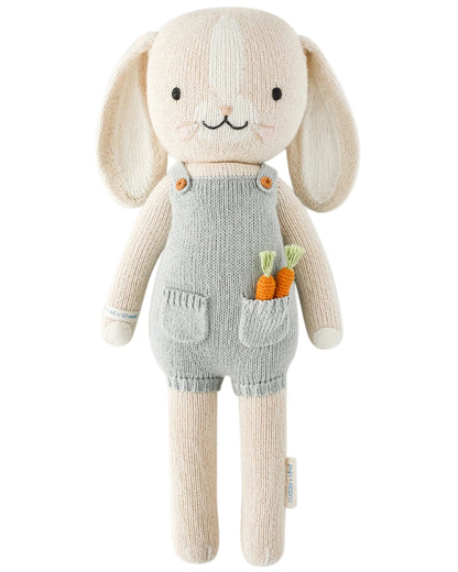 Henry the Bunny - Little 13" by Cuddle + Kind is a knitted beige bunny in grey overalls with two carrots in the pocket—perfect for a little carrot connoisseur—posed against a white background.