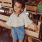 A young girl in a Louise Misha Girls' Feliza Blouse and denim shorts stands by a wooden vegetable display at an outdoor market.