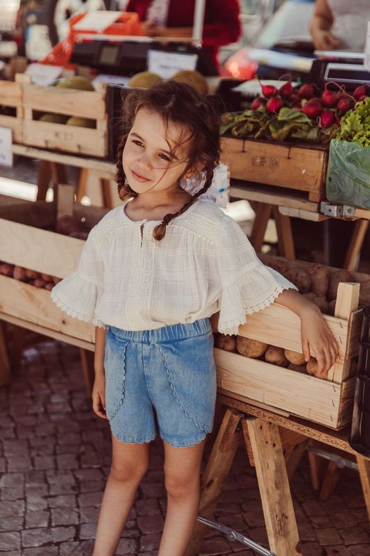 A young girl in a Louise Misha Girls' Feliza Blouse and denim shorts stands by a wooden vegetable display at an outdoor market.