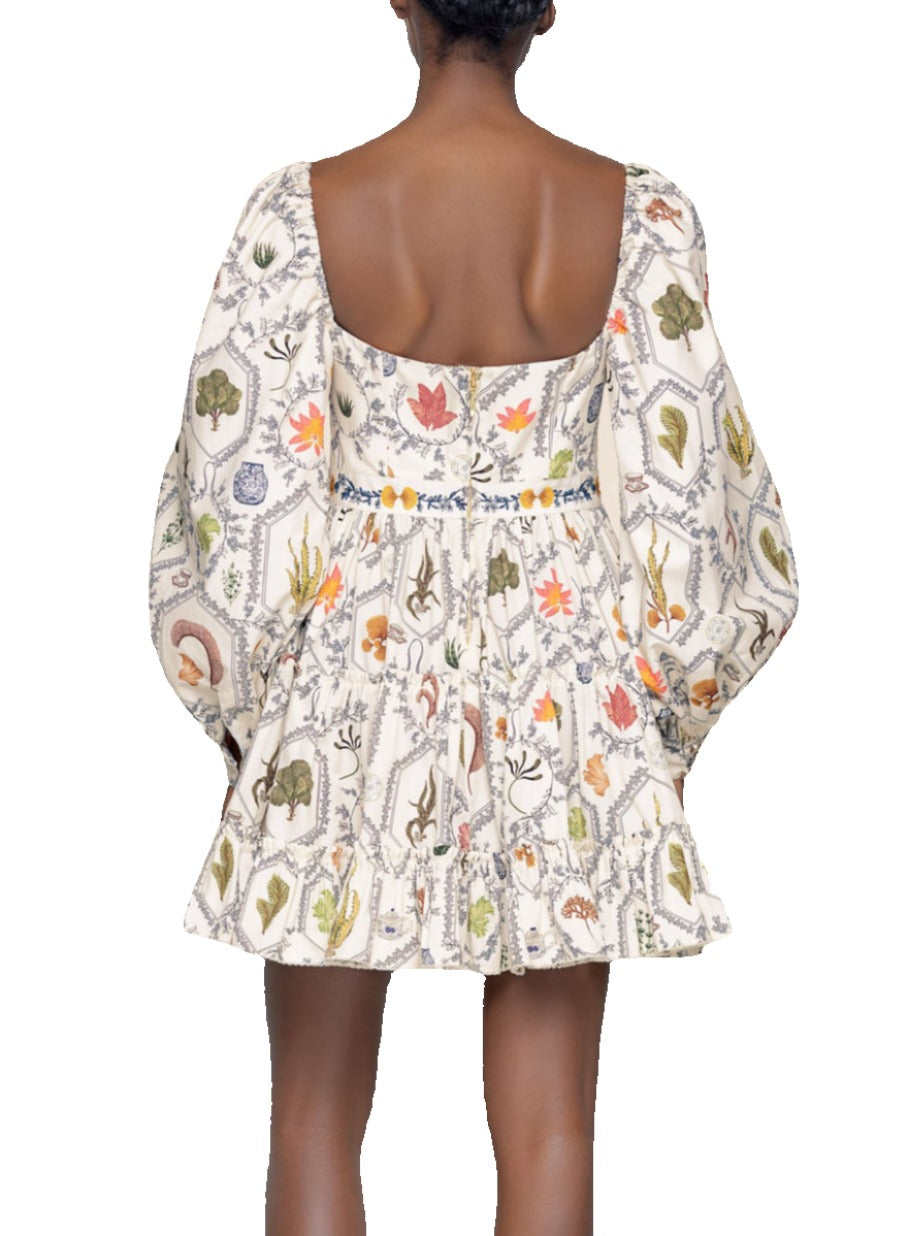 Rear view of a woman wearing a short, white Agua by Aguabendita Honor Dress mini dress with colorful botanical prints, featuring a low-cut back and balloon sleeves.