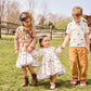 Three children holding hands and walking in a grassy area, with two girls wearing Pink Chicken Girls' Julia Dresses with chambray detailed tiers and a boy sporting an equestrian chic polo shirt and pants. Trees and a wooden fence are visible in the background.