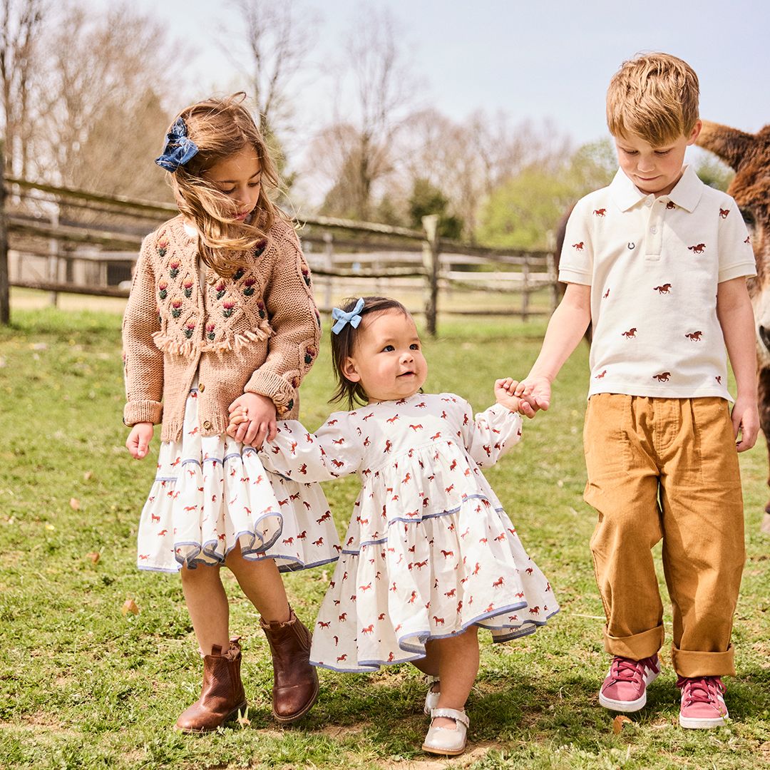 Three children holding hands and walking in a grassy area, with two girls wearing Pink Chicken Girls' Julia Dresses with chambray detailed tiers and a boy sporting an equestrian chic polo shirt and pants. Trees and a wooden fence are visible in the background.