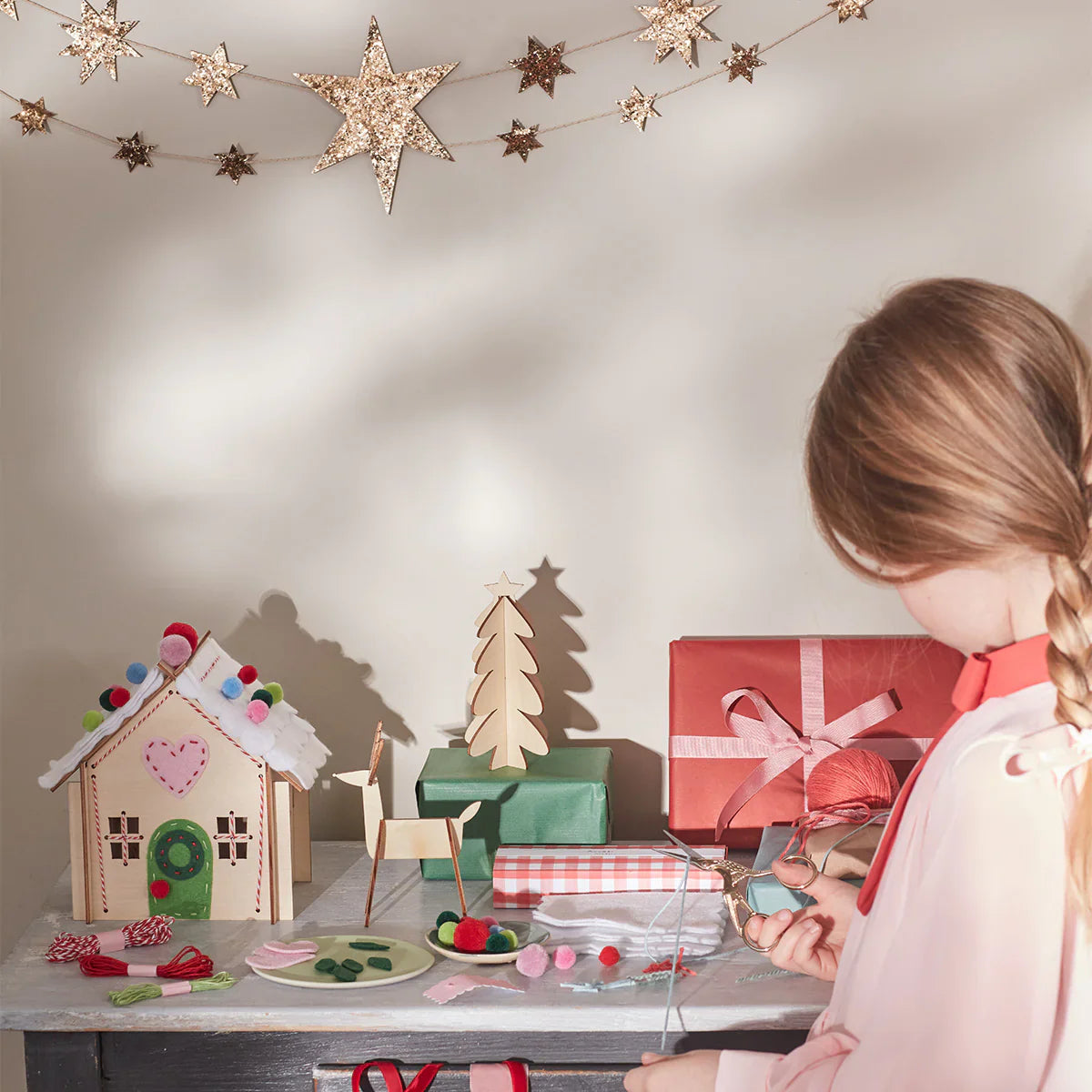 A creative child with braided hair is decorating a table with holiday-themed crafts, including a gingerbread house and wrapped gifts. Star-shaped decorations hang above, alongside the Meri Meri Wooden Embroidery Gingerbread House Kit, which makes for the perfect Christmas gift.