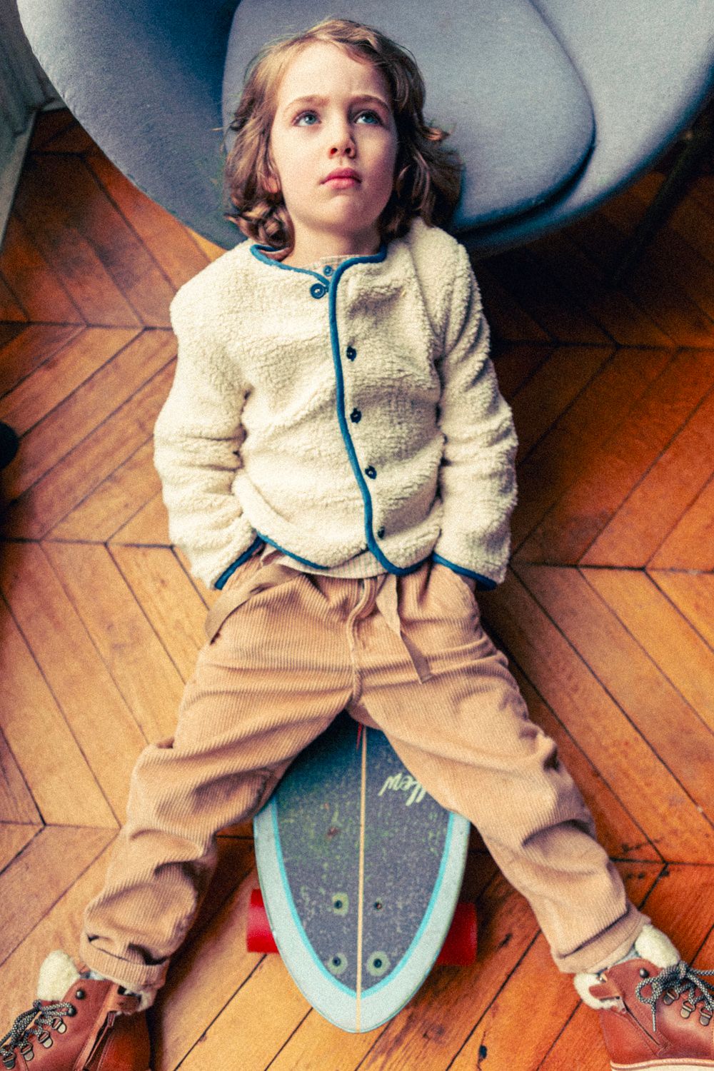 A child with wavy hair sits on a skateboard indoors, leaning back against a chair, wearing the Louis Louise Baby Boys' and Boys' Teddy Bear Fake Fur Jacket in cream with tan pants.