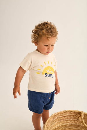A young child with curly hair wears the Cozmo Baby Boys' Ian T-Shirt, featuring a yellow sun and "Yes, Sun" text, paired with blue shorts. He stands near a woven basket on a plain background.