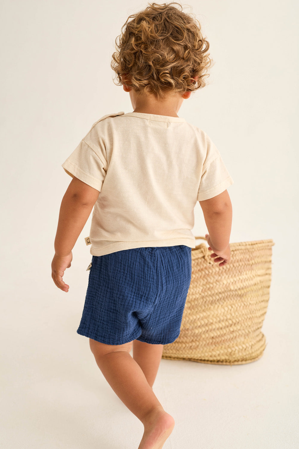 A toddler with curly hair stands barefoot near a woven basket, facing away from the camera in a Cozmo Baby Boys' Ian T-Shirt and blue shorts.
