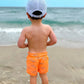 A young child in orange shark-print shorts and a Bits & Bows Lil Bro Baseball Hat stands barefoot on the beach, gazing at the ocean.