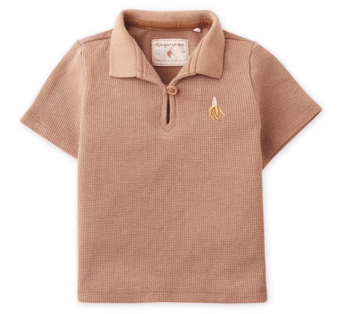 The Gingersnaps Baby Boys' Timmy Waffle Knit Polo Shirt features short sleeves, a light brown color, a collar, and a small embroidered banana logo on the chest—perfect for stylish baby wear.