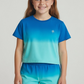 A girl stands facing forward, smiling with long brown hair, wearing Feather 4 Arrow’s Baby Girls’ and Girls’ Weekend Short—a pair of matching French terry sweatshorts with an elastic waistband and drawcord—paired with a blue and turquoise gradient t-shirt.