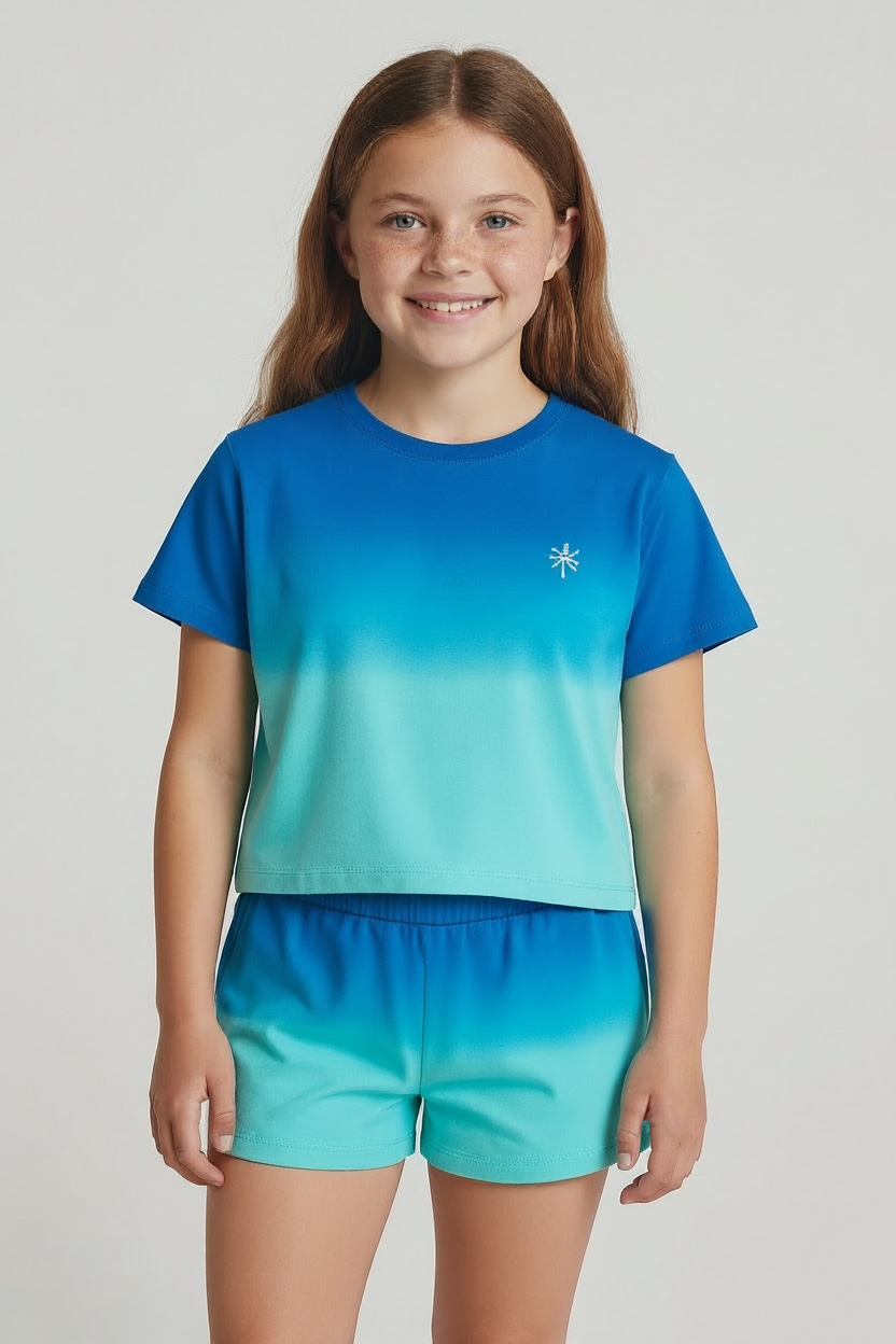 A girl stands facing forward, smiling with long brown hair, wearing Feather 4 Arrow’s Baby Girls’ and Girls’ Weekend Short—a pair of matching French terry sweatshorts with an elastic waistband and drawcord—paired with a blue and turquoise gradient t-shirt.