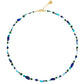 The Milos Beaded Necklace by Anabel Aram features small blue, turquoise, green, and gold beads with a gold clasp and adjustable chain.