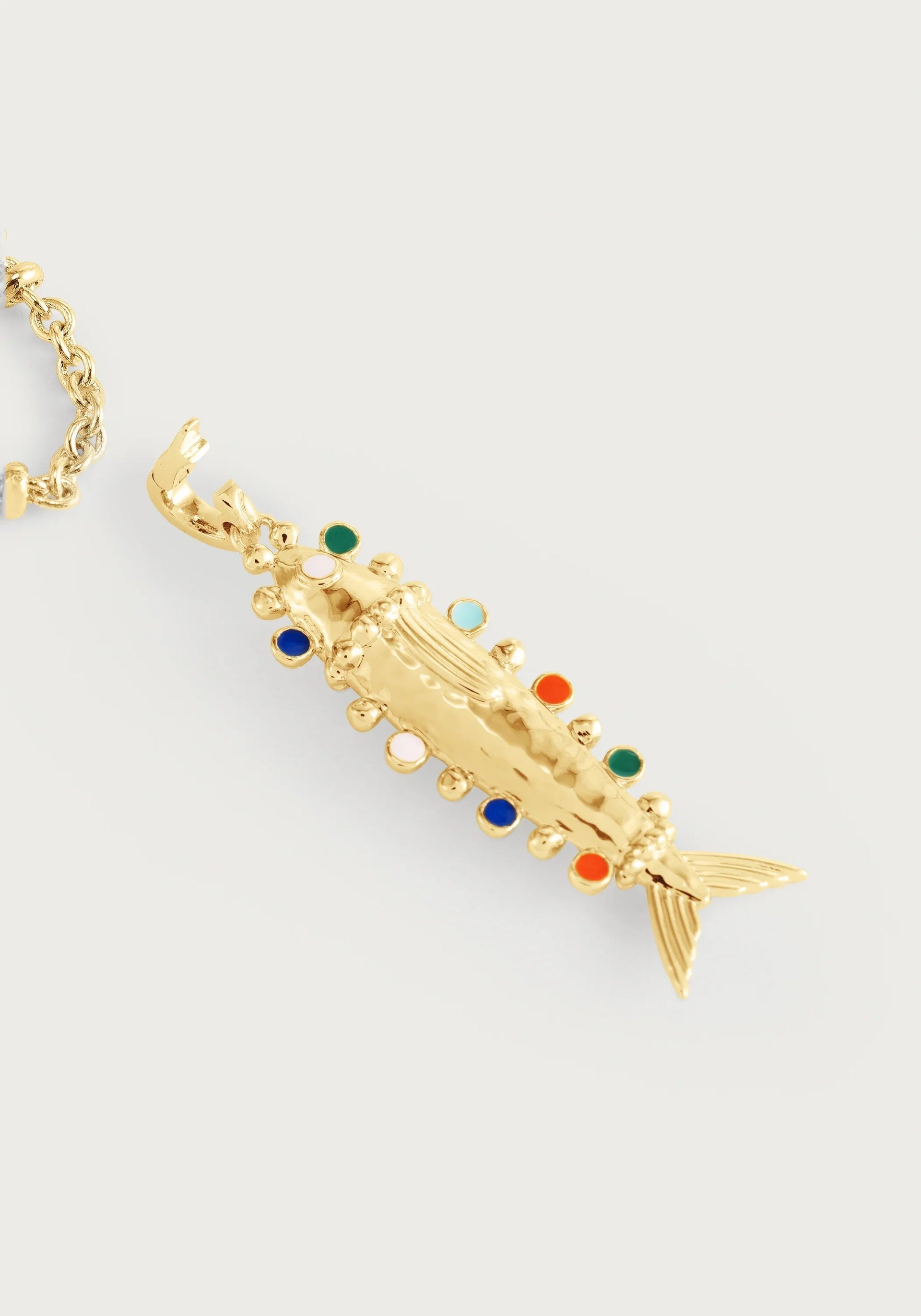 The Milos Medium Fish Charm by Anabel Aram is a gold pendant with textured details and colorful gemstone accents, capturing Mediterranean style. Shown on a gold necklace against a light background.
