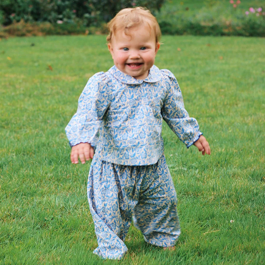 A smiling toddler with curly hair stands on grass, wearing the Pink Chicken Baby Girls' Dawson 2-Piece Set in a blue climbing floral print, made from soft cotton poplin.