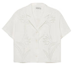 The Louisa Camp Shirt by Simkhai is a white short-sleeve top featuring a notched collar and subtle embroidered floral patterns on the front.