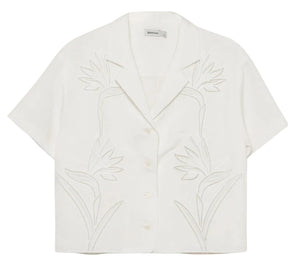 The Louisa Camp Shirt by Simkhai is a white short-sleeve top featuring a notched collar and subtle embroidered floral patterns on the front.