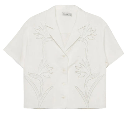 The Louisa Camp Shirt by Simkhai is a white short-sleeve top featuring a notched collar and subtle embroidered floral patterns on the front.