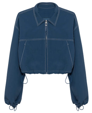 The Colsen Cotton Cropped Jacket by Simkhai features dark indigo cotton, white contrast stitching, long sleeves, a collar, and adjustable drawstrings at the hem and cuffs.