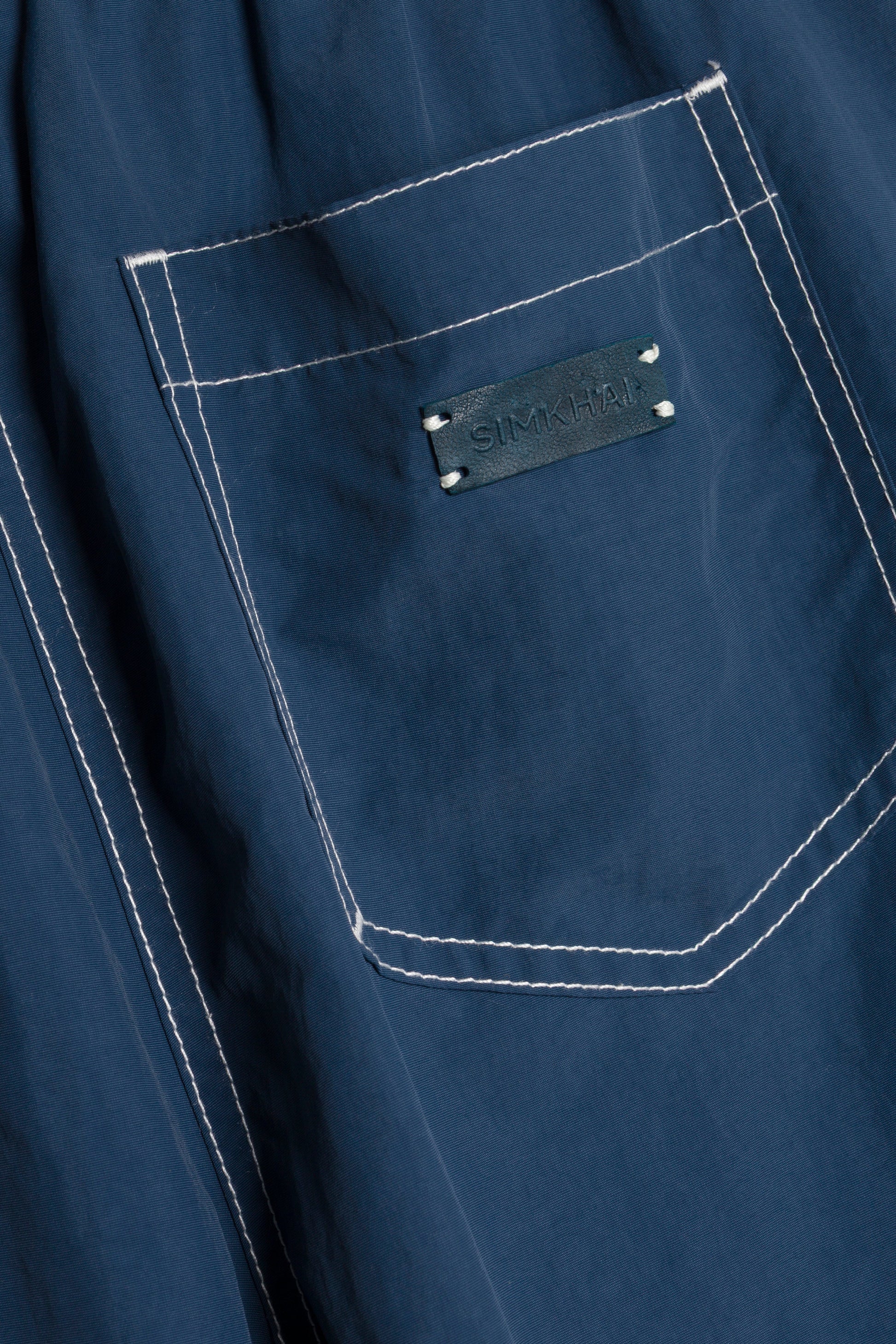 Close-up of a blue fabric pocket with white stitching and a black SIMKHAI label above the pocket, featured on the Lennox Cotton Straight Leg Pant by Simkhai.