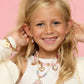 A young girl with long blonde hair smiles in front of a pink background, wearing Super Smalls pastel jewelry and the Cupcake Lipgloss Locket pendant.